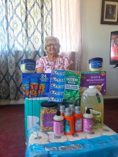 Hopi elder receives a coronavirus emergency hygiene and food supply box shipped by Crossing Worlds Hopi Projects. She has displayed the toilet paper, cleaning, toiletry and food items in front of her.