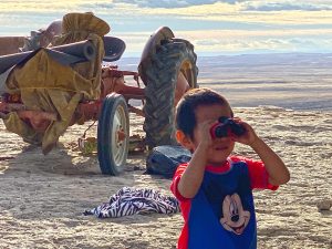 Hopi boy with new binoculars on top of cliff edge with tractor in background represents Hopi youth looking to the future and welcoming learning enrichment supplies and vision for the future