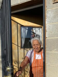 Hopi elder woman in apron looking out from open door of block house.